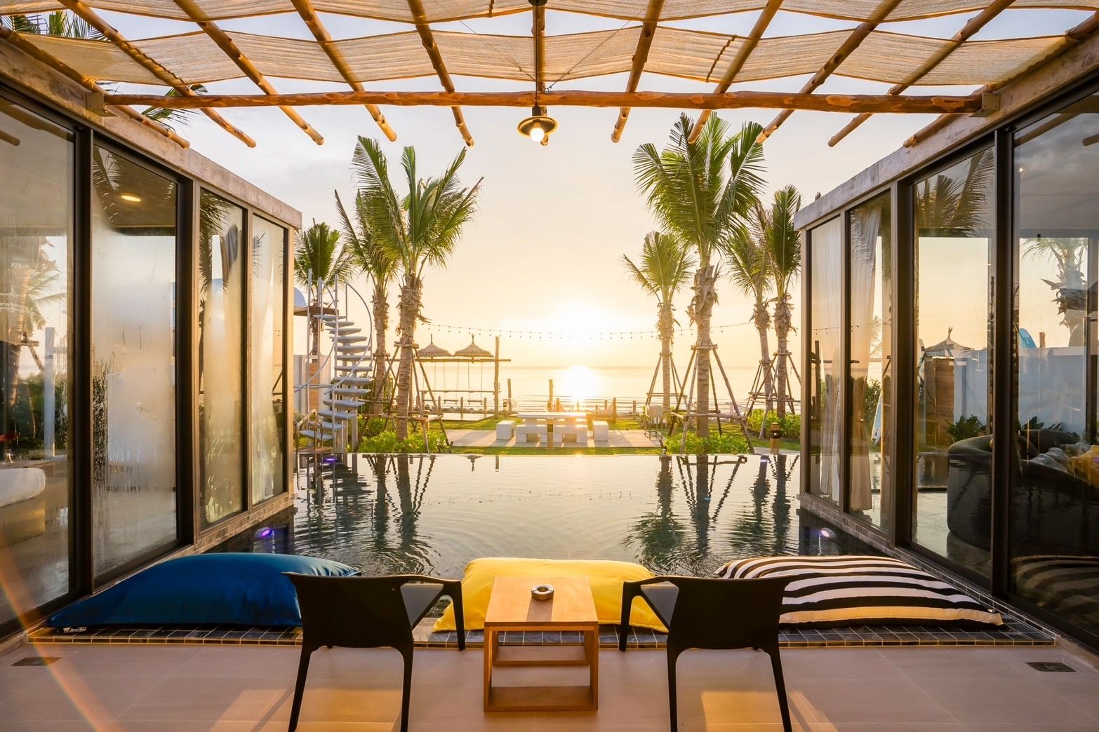 Sunset view from Amore' A Pool Villa, หาดปึกเตียน-ชะอำ. Infinity pool reflects palm trees and golden sky. Chairs and cushions