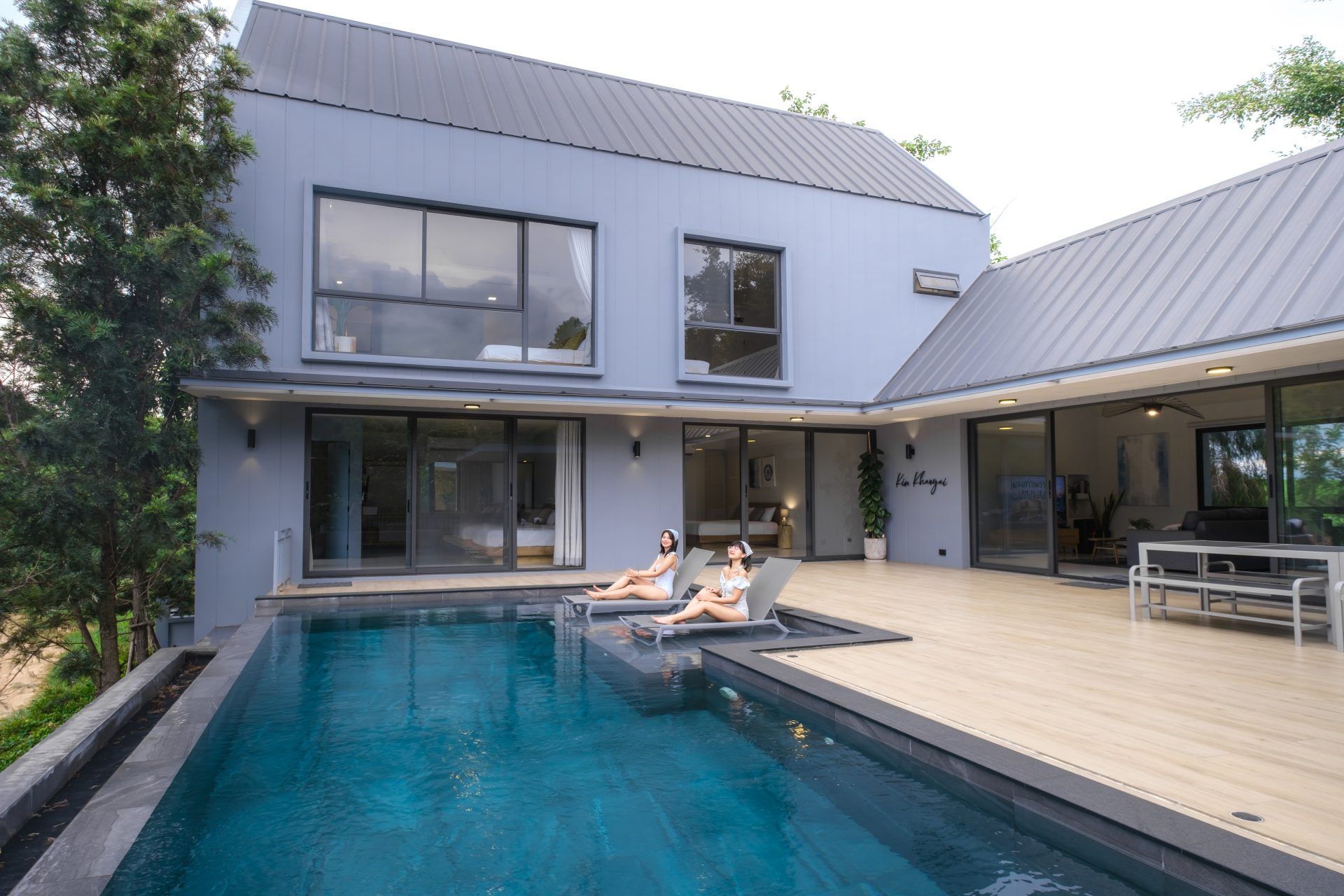Two women relax on lounge chairs by the pool at Kin Khaoyai V Pool Villa, หมูสี เขาใหญ่. Modern villa exterior.