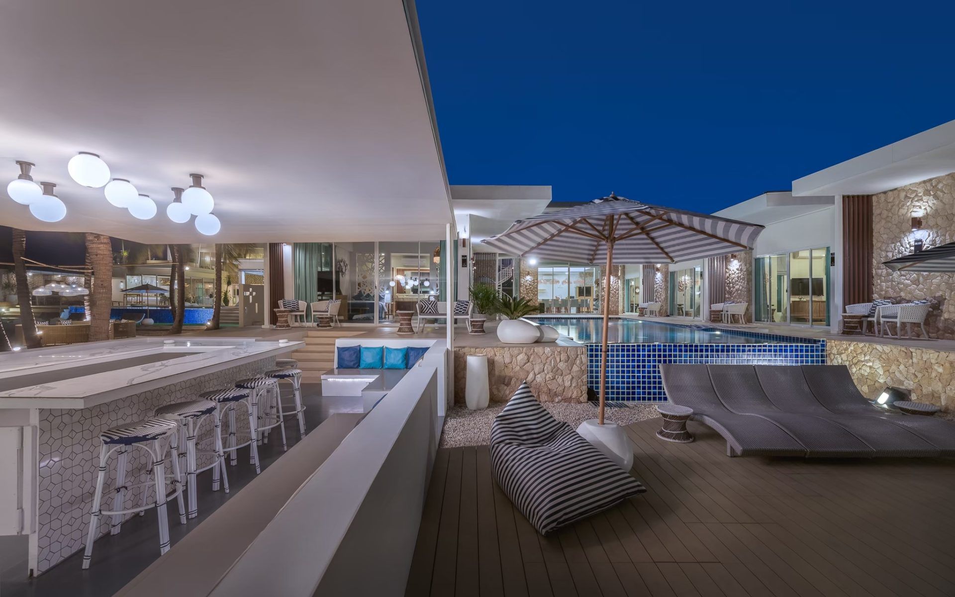 Evening view of Vela VI pool villa in หาดปึกเตียน ชะอำ, Thailand. Features poolside bar, seating, and striped umbrella.