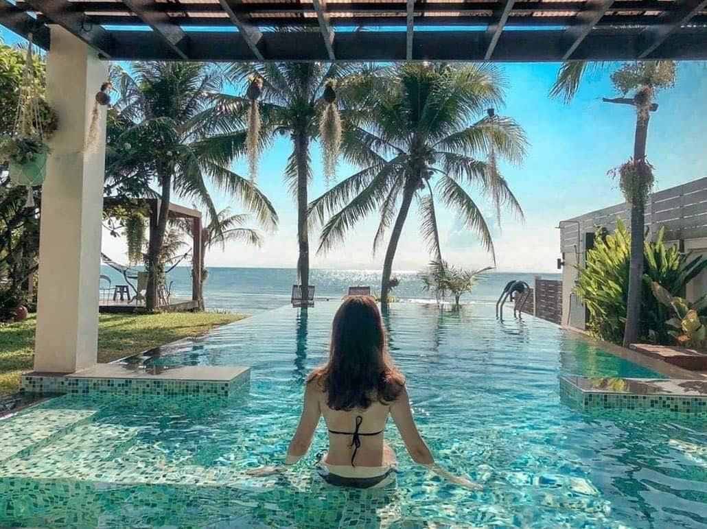 Woman enjoys the infinity pool overlooking the ocean at Pran Beach Villa I, เขากะโหลก ปราณบุรี. Palm trees and blue sky.