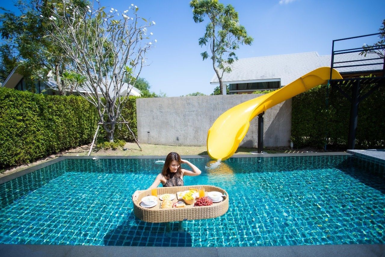 Woman enjoys floating breakfast in the pool with waterslide at Proud IV Pool Villa, Hua Hin, Thailand.