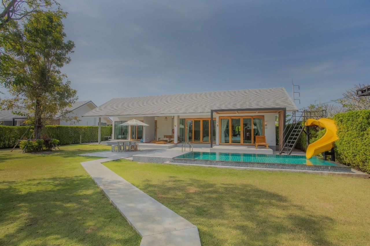 Daytime view of the pool and slide at Proud V Pool Villa, Hua Hin. Lush green lawn and modern villa design.