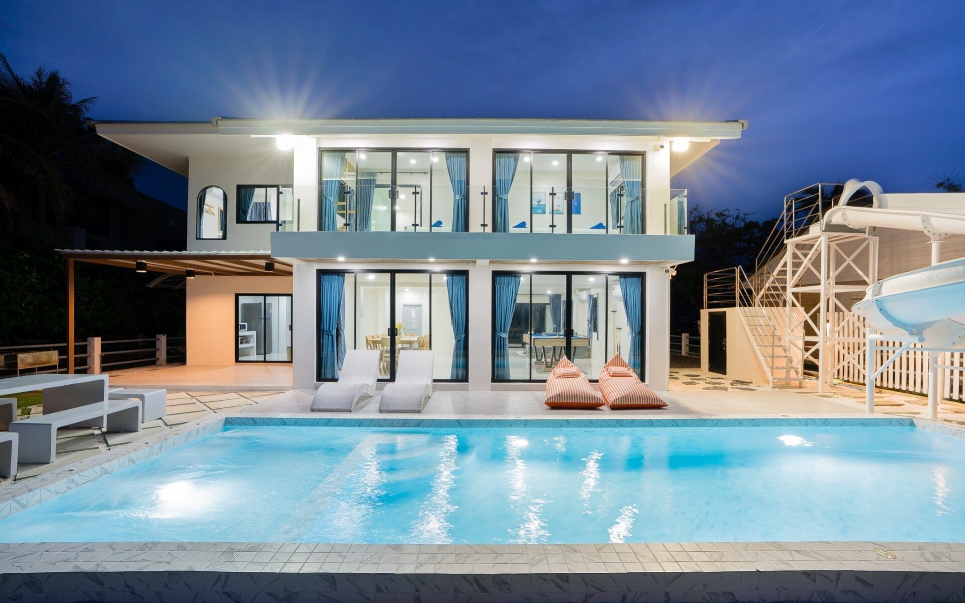 Night view of the pool and villa exterior at Sea Time V Pool Villa, Cha-Am. Features a waterslide and lounge chairs.