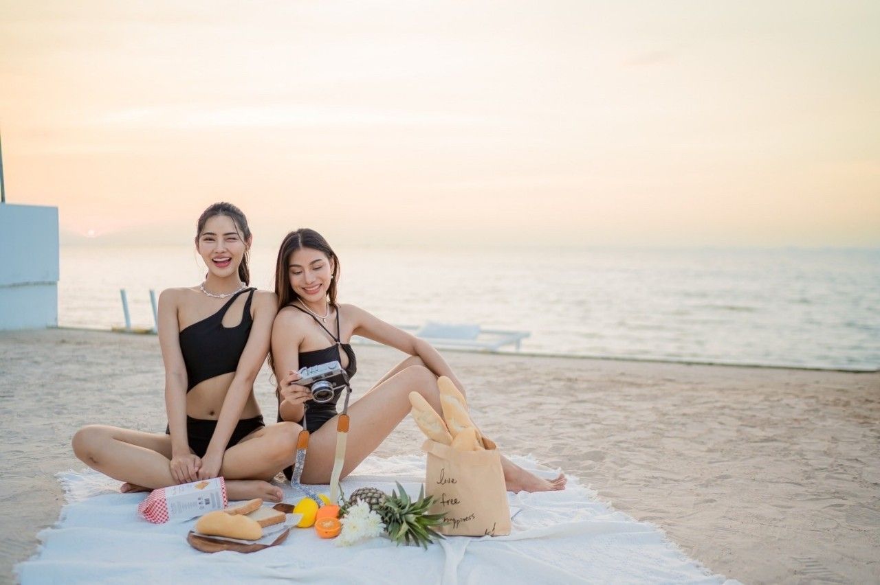 Two women enjoy a beach picnic at Sea View IV Pool Villa, พัทยาเหนือ- บางละมุง. One holds a vintage camera.