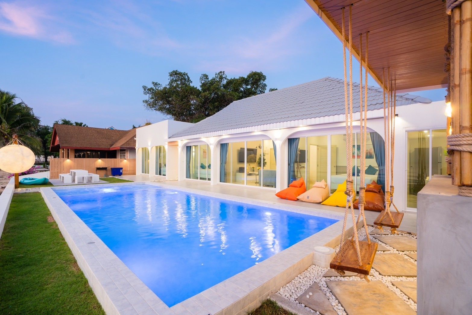 Sun Sea V Pool Villa in Cha Am features an illuminated pool and swings. Bean bag chairs line the villa's edge.