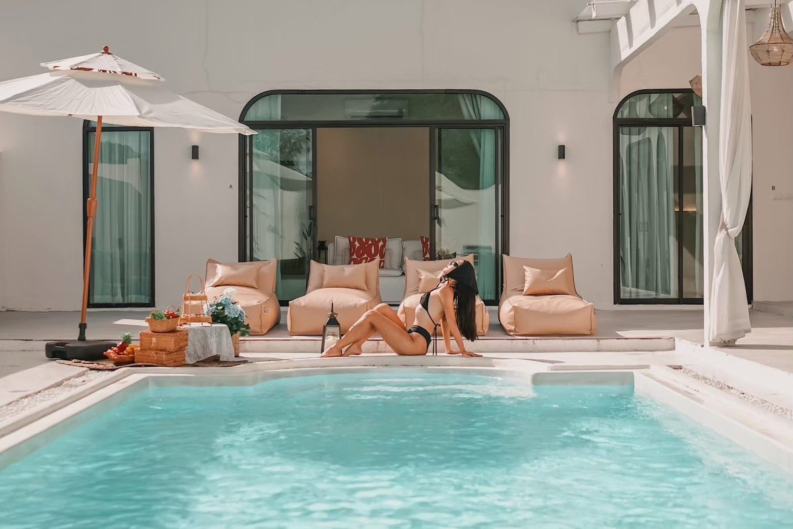 Woman relaxes by the pool at White Coco V Pool Villa, พัทยากลาง-นาเกลือ. View of lounge chairs and bedroom through sliding gl