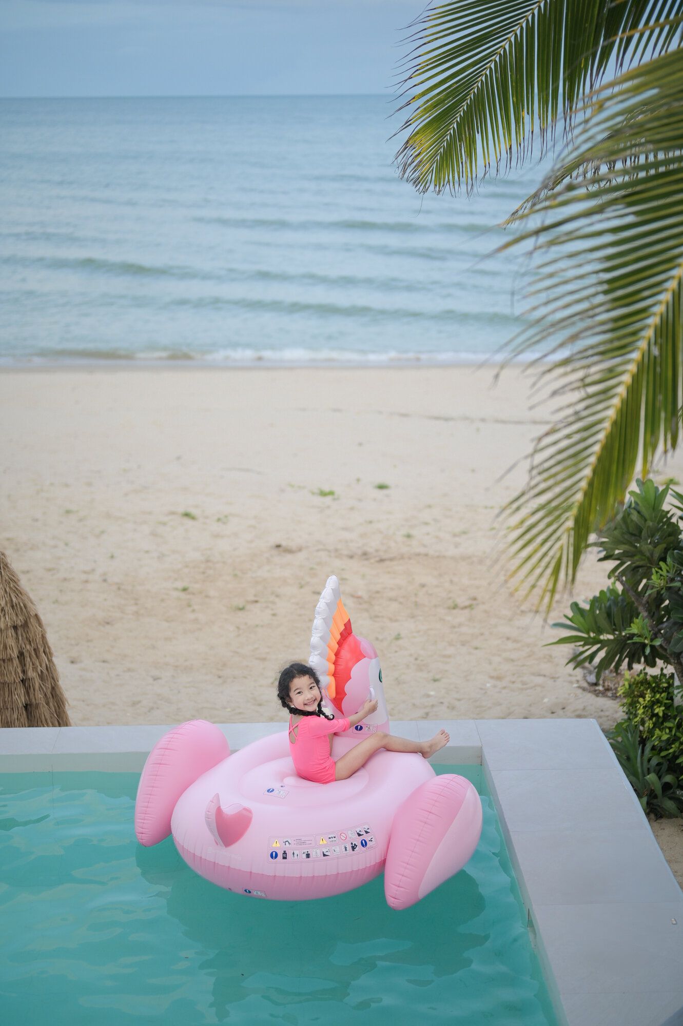 A child on a pink unicorn float in the pool at Coastal Calm IV Pool Villa, Cha-Am, with the beach and ocean in the backgroun
