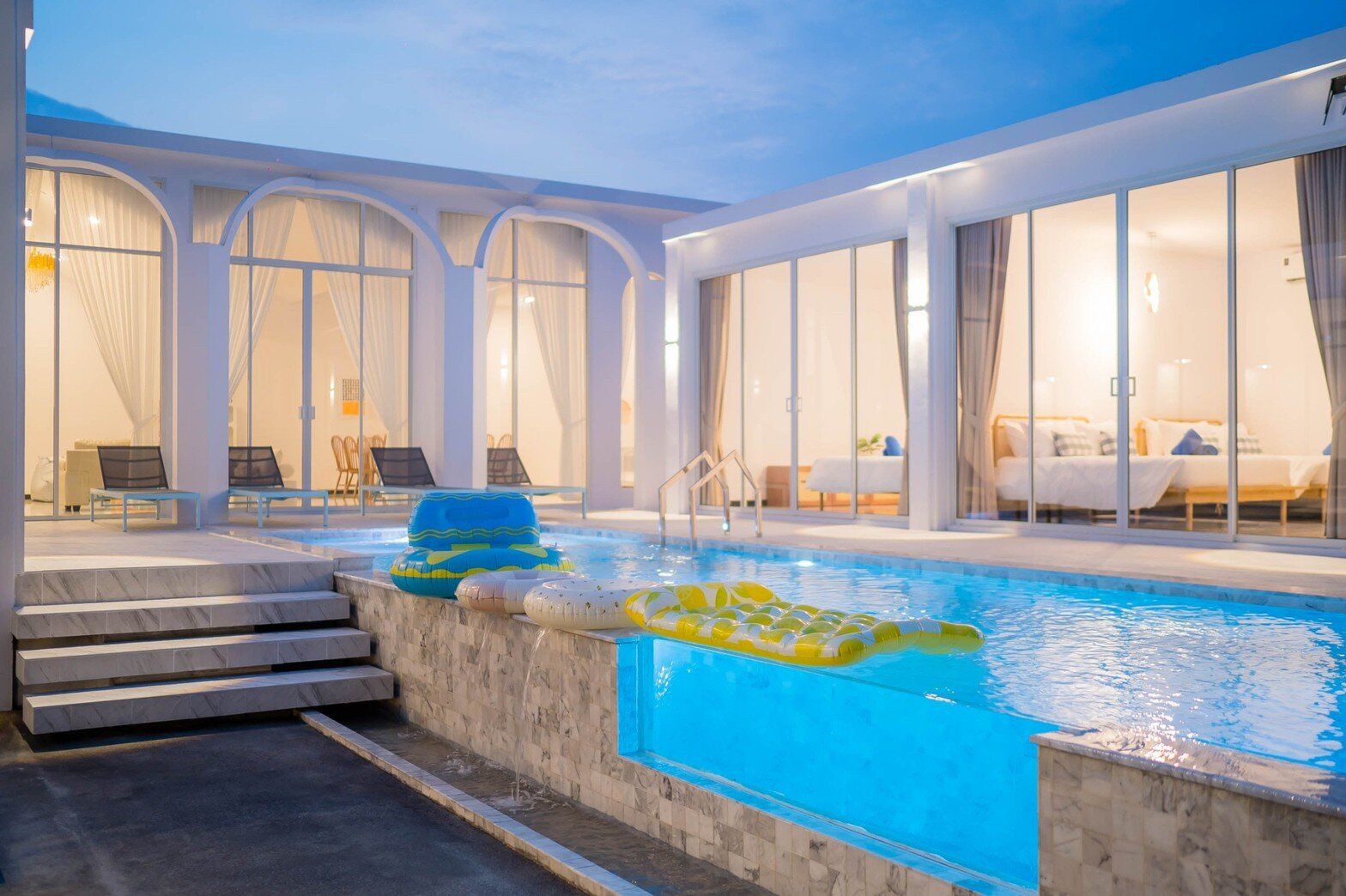Miami Beach VI Pool Villa in Cha-Am features a stunning glass-wall pool with vibrant blue lighting and inflatables.