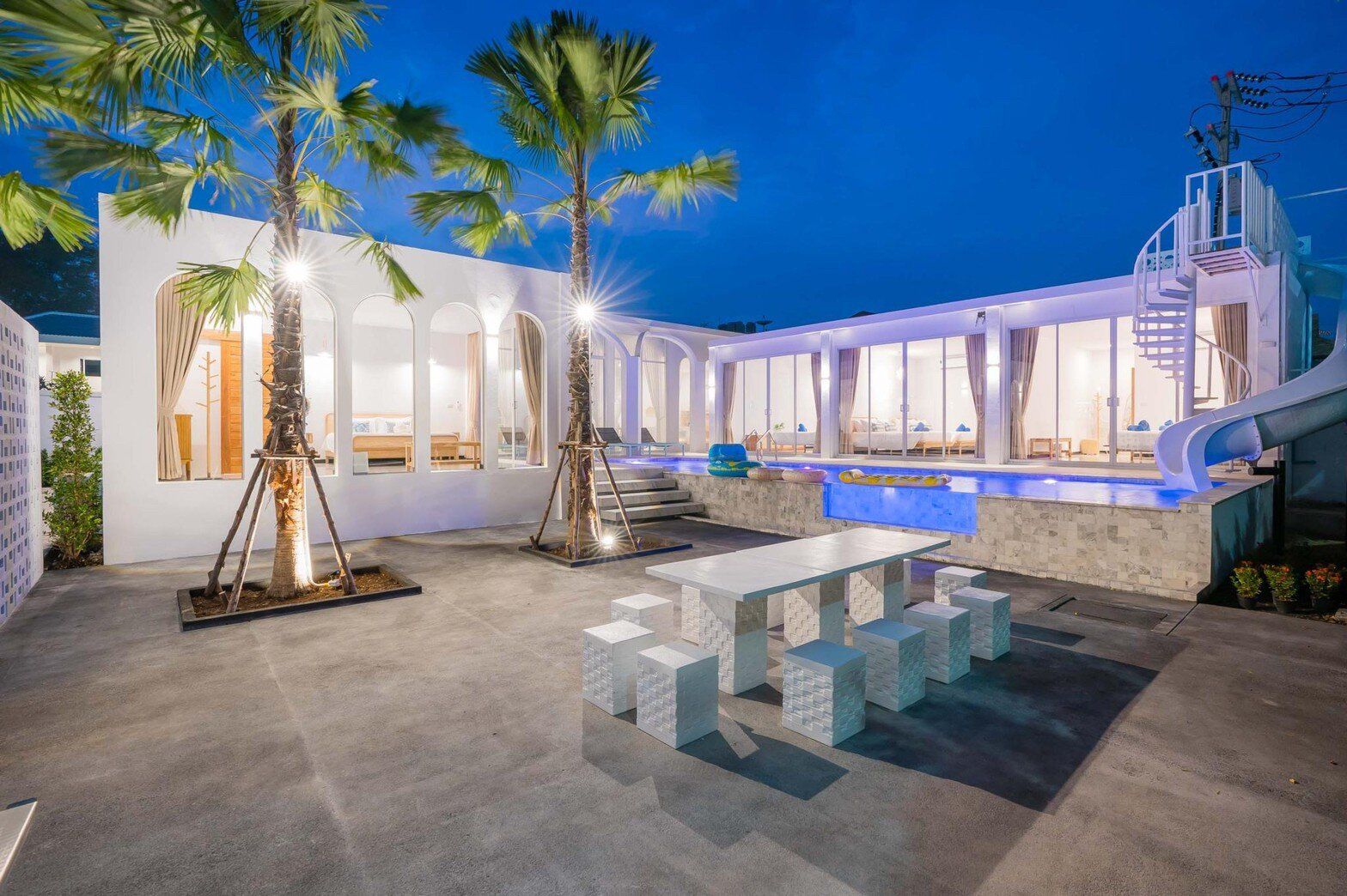 Miami Beach VI Pool Villa in Cha-Am at dusk, featuring a bright blue lit pool with a waterslide and spiral staircase.