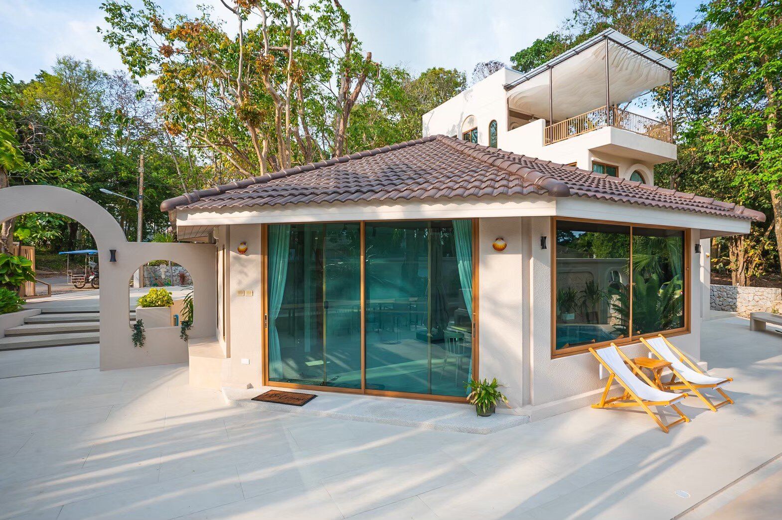 Daylight view of Sand Moment VI Pool Villa entrance in Rayong, featuring arched walls, tiled roof, and lush greenery.