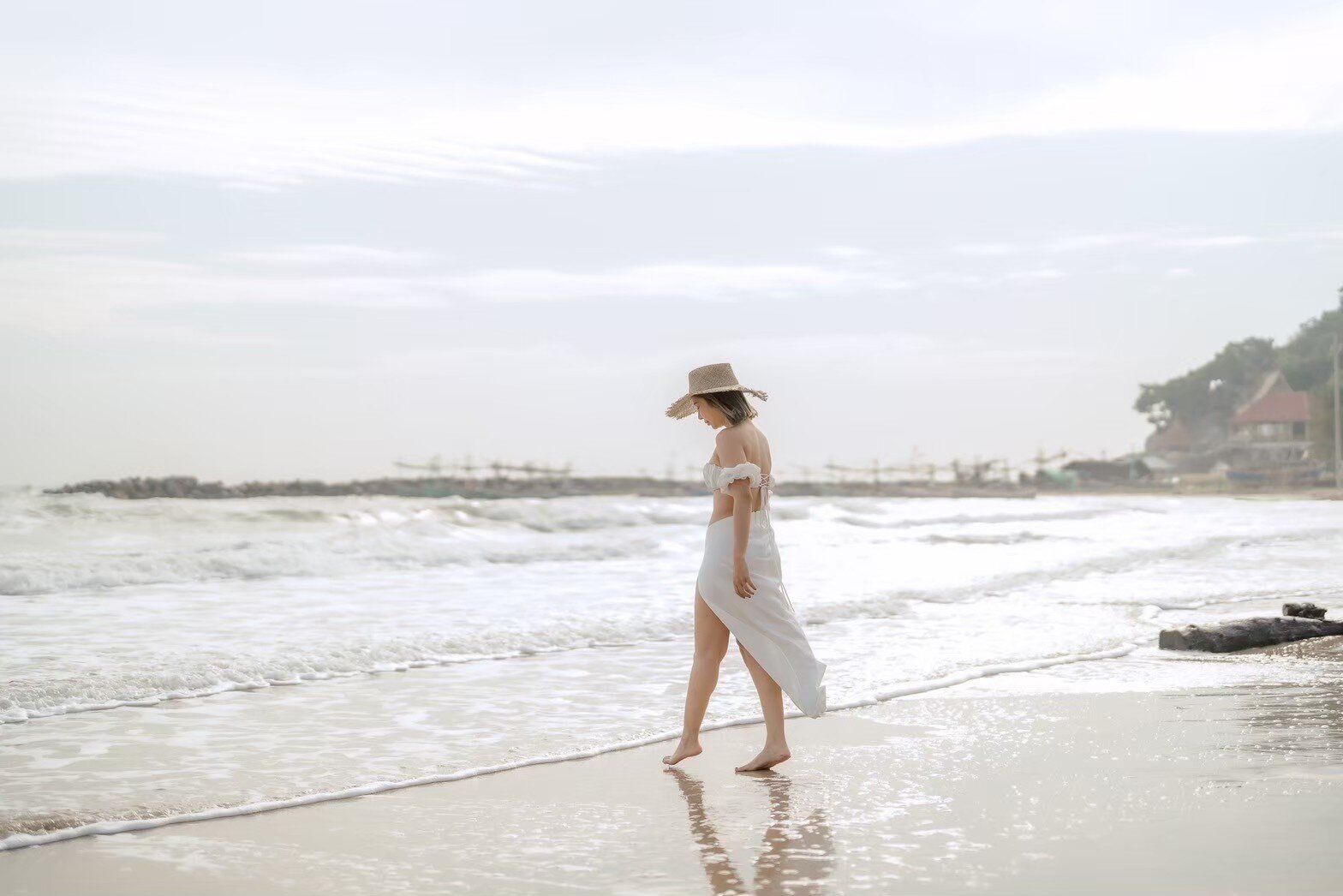 A woman in a straw hat and white dress walks along the sandy beach with waves at Story II Pool Villa Hua-Hin.
