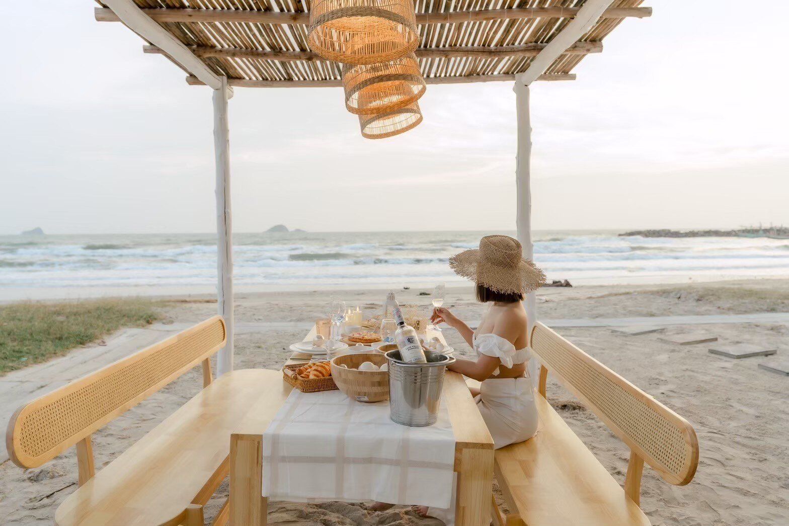 A woman enjoys beachfront dining under a bamboo canopy at Story II Pool Villa Hua-Hin, with the ocean in the background.