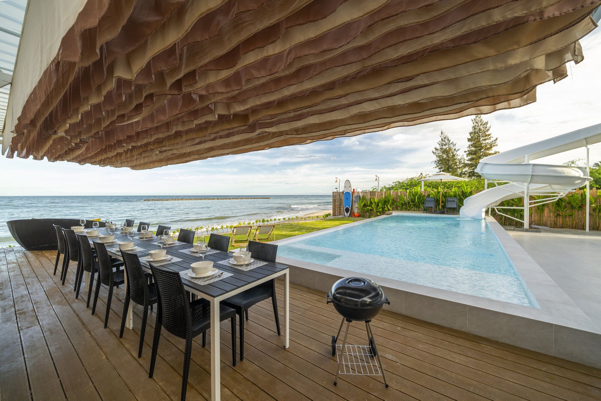 Beachfront outdoor dining table set for guests at Talay Haus X Pool Villa in Cha-Am, with a BBQ grill and pool slide.
