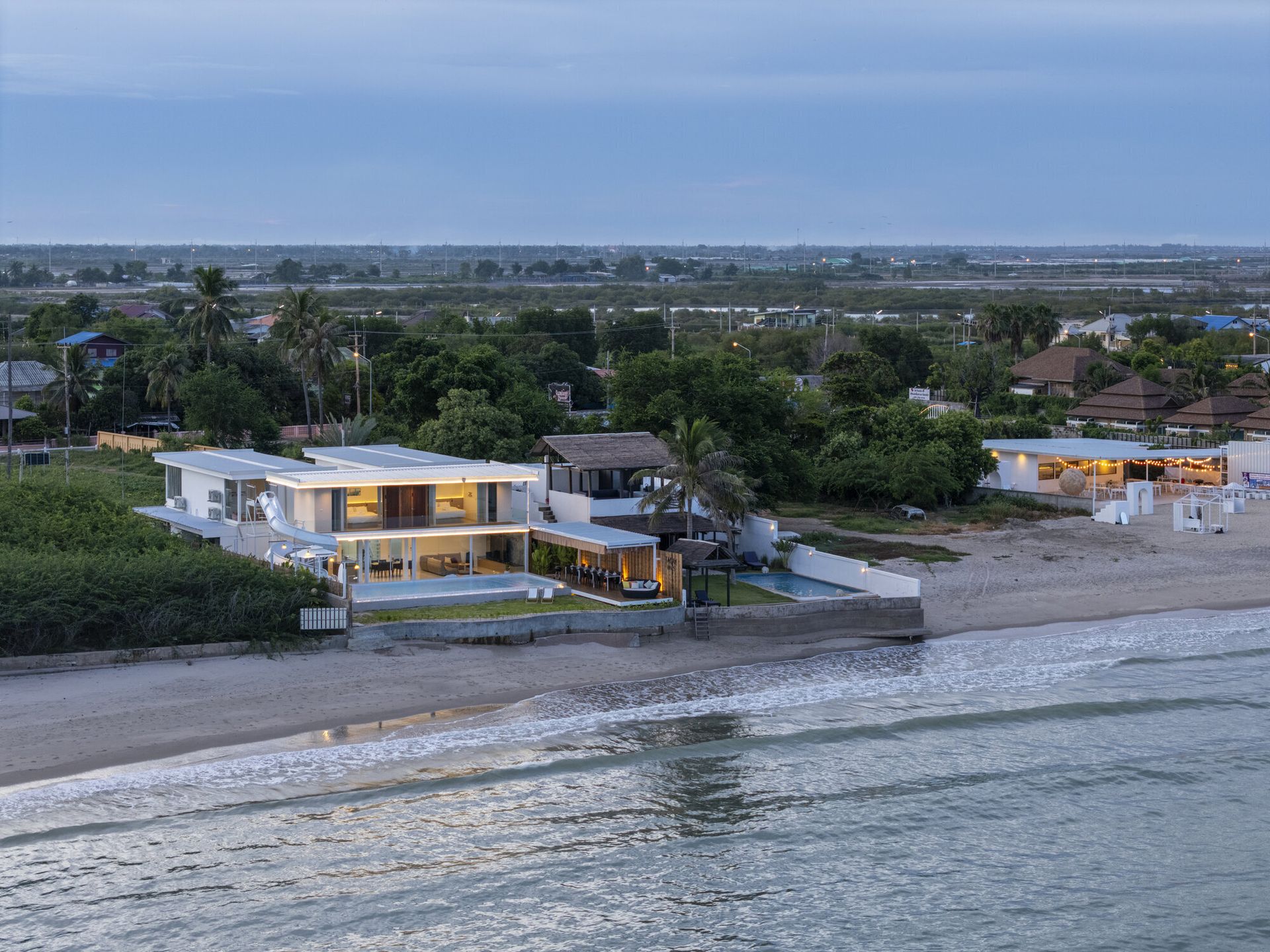 Aerial view of Talay Haus X Pool Villa in Cha-Am at dusk, showcasing its modern design, beachfront location, and illuminated interiors.
