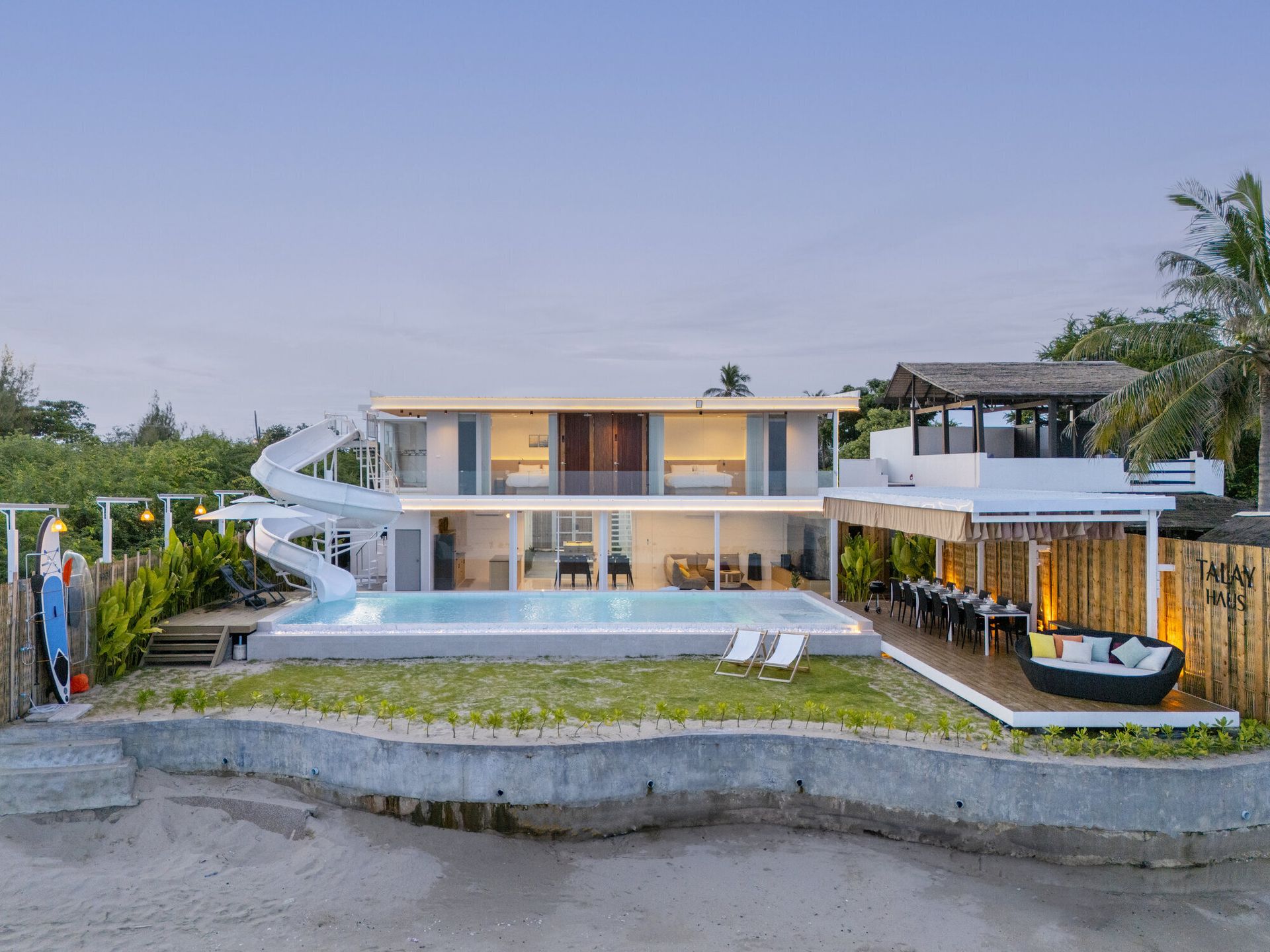 Aerial view of Talay Haus X Pool Villa in Cha-Am, showing the modern beachfront villa, pool, water slide, and outdoor dining area at dusk.