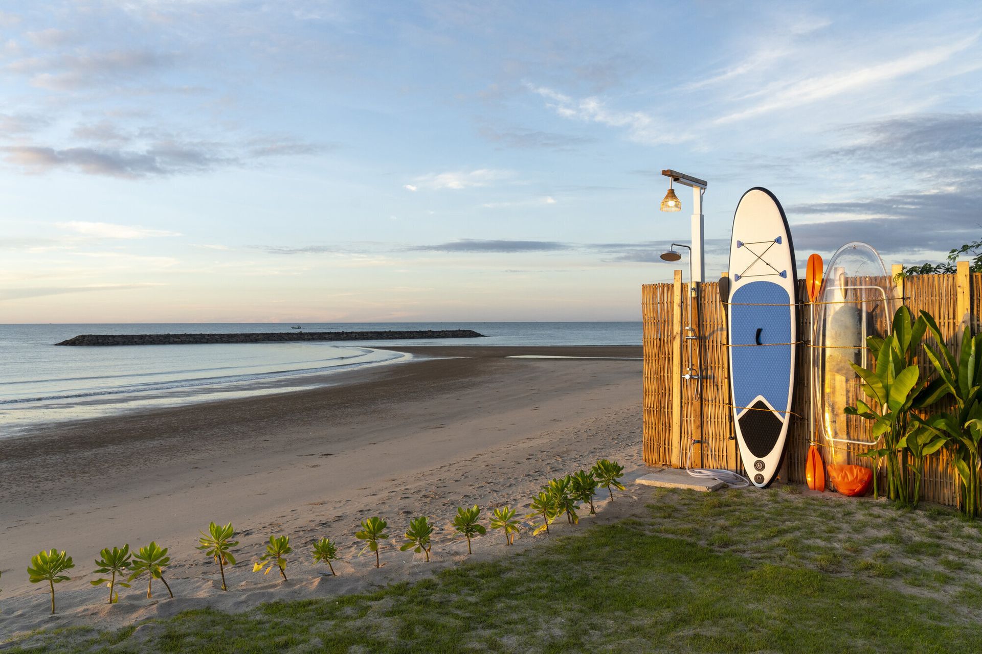 Beachfront outdoor shower and paddleboards at Talay Haus X Pool Villa in Cha-Am, with the ocean and sandy beach at sunset.