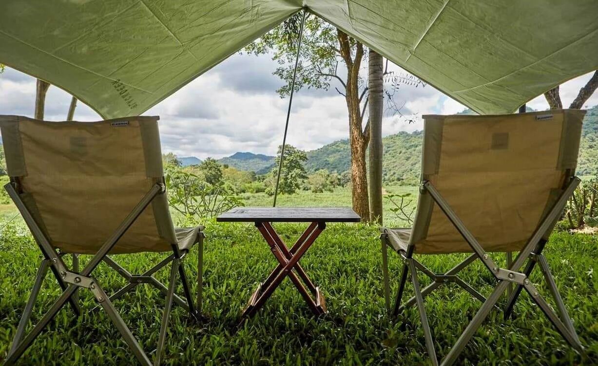 Two camping chairs and a small table under a tarp, facing lush green mountains at The Valley Khaoyai I, Khao Yai.