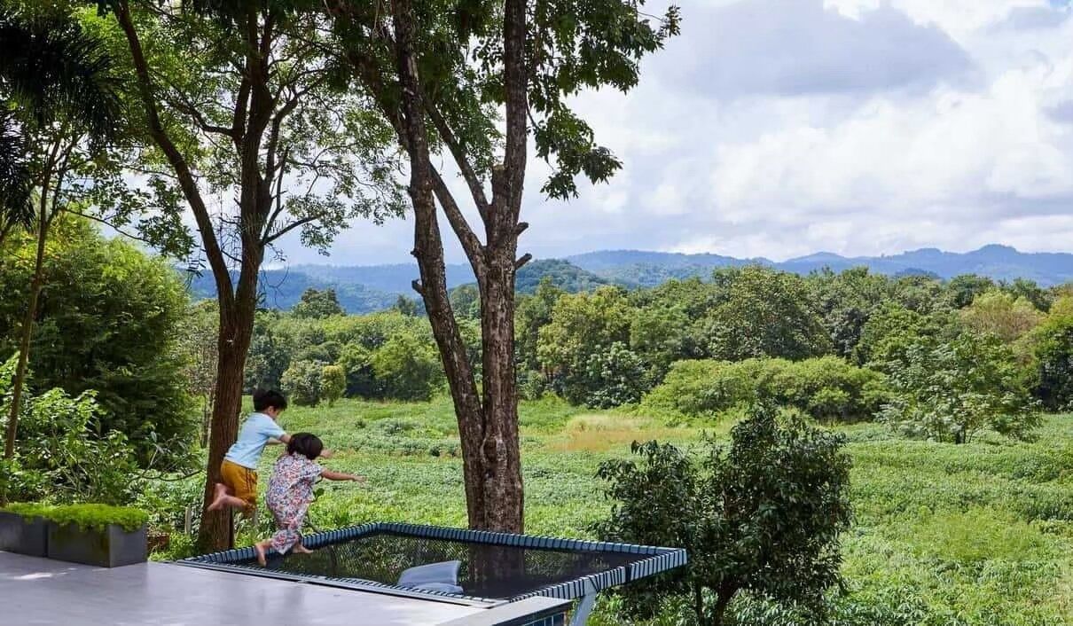 Two children jumping on an outdoor trampoline at The Valley Khaoyai I, with lush forest and mountains in the background.