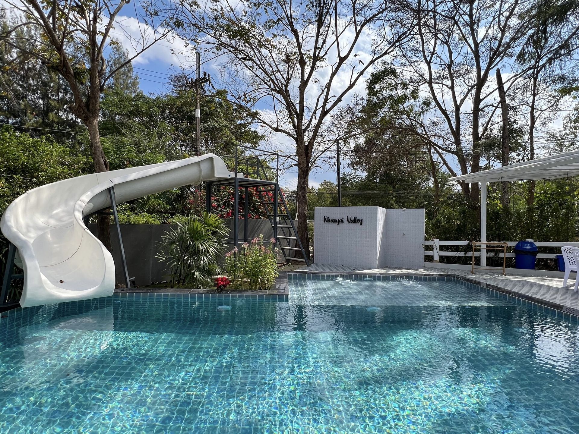 The Valley Khaoyai IX Villa's outdoor pool with a white water slide, surrounded by lush greenery under a blue sky.