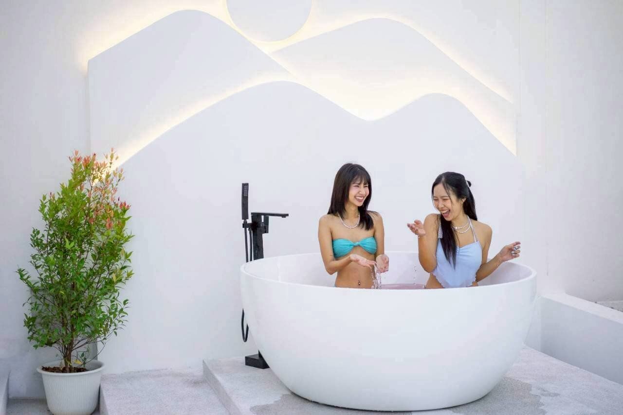 Two women laughing in a large outdoor bathtub at Tierra V Pool Villa, Pattaya, with modern white wall decor and a plant.