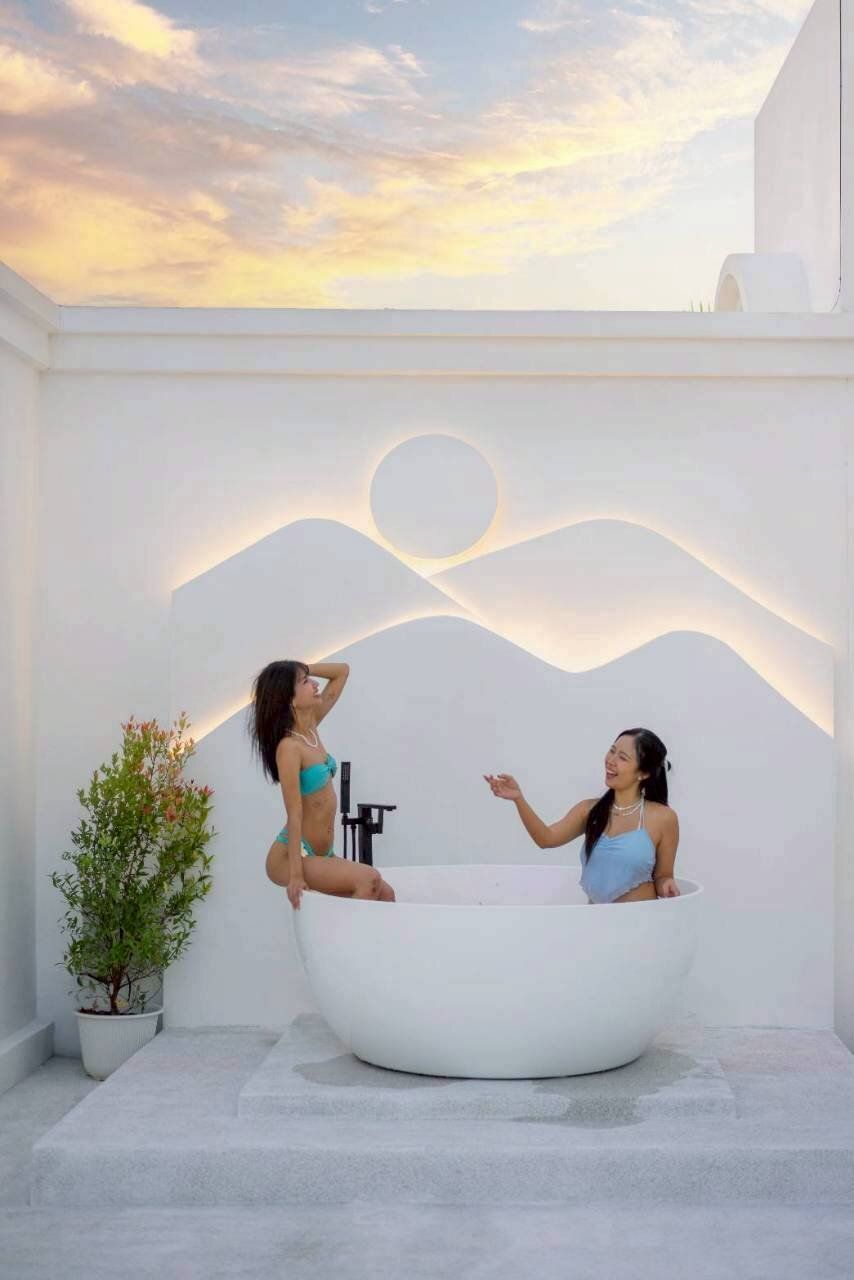 Two women enjoying an outdoor bathtub at Tierra V Pool Villa, Pattaya, with a beautiful sunset sky and unique wall art.
