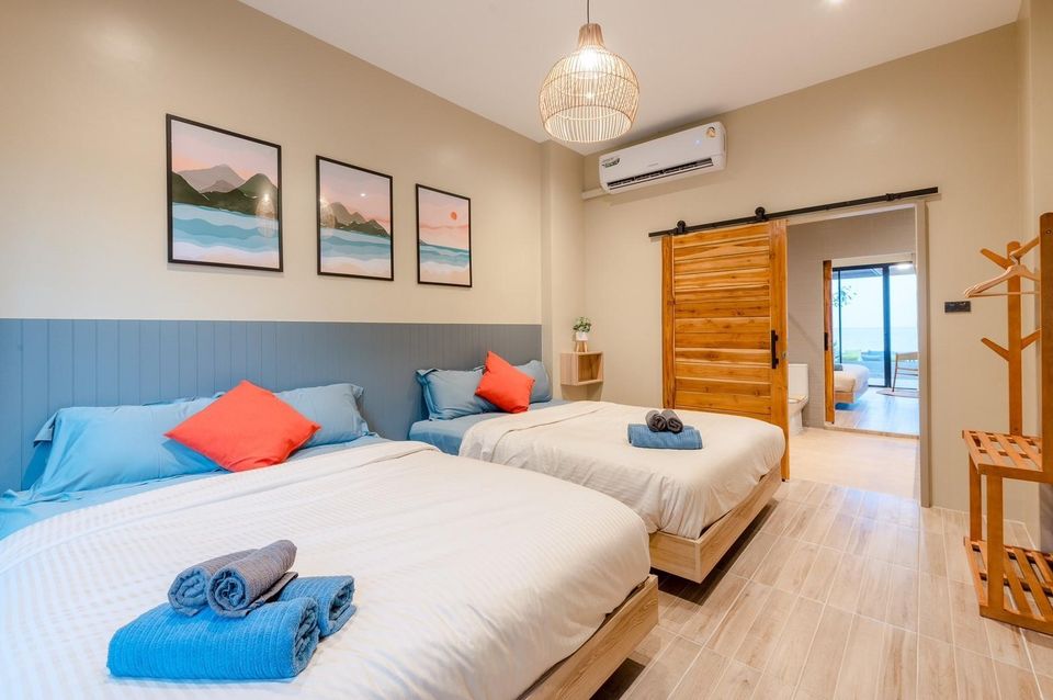 Bright bedroom at Beach Blanca IV Pool Villa, Cha-Am, featuring two comfortable beds with blue linens and orange pillows. Sliding door leads to a balcony.