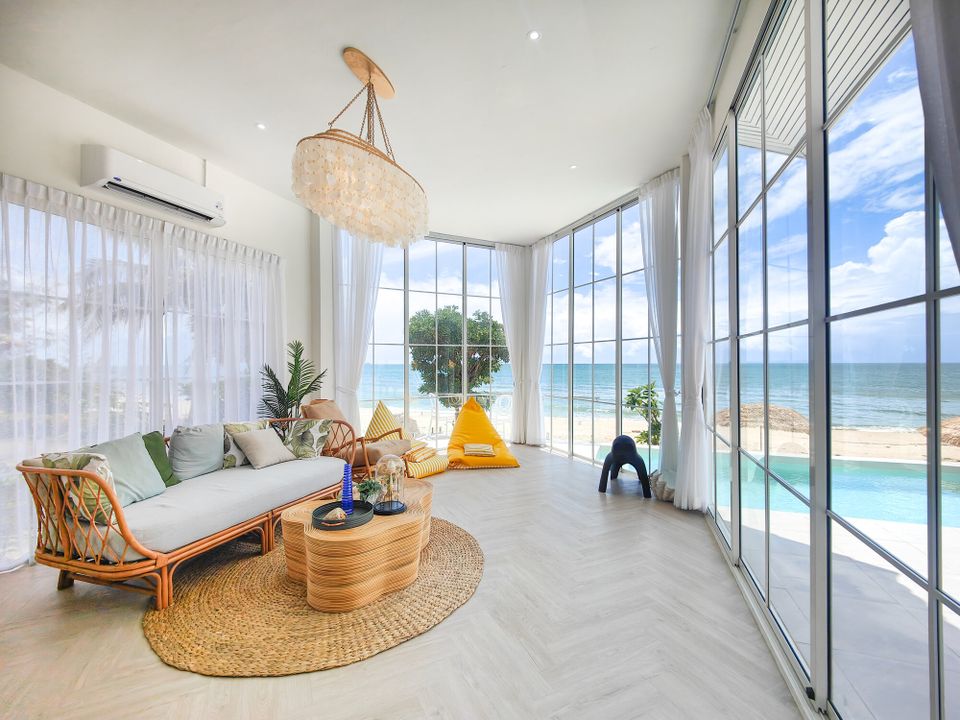 Bright beachfront living room at Coastal Calm IV Pool Villa, Cha-Am, with rattan sofa, shell chandelier, and ocean views.