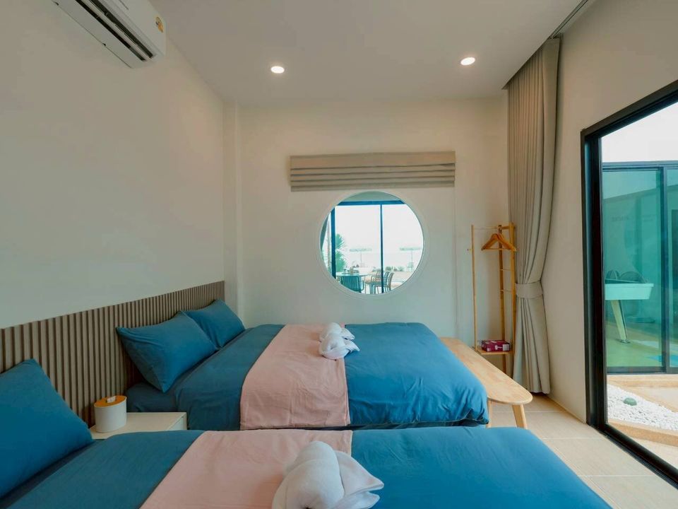 Bright bedroom at Darlington IV Pool Villa, Cha-Am, with two twin beds, blue and pink bedding, and a unique porthole window overlooking the outdoor area.
