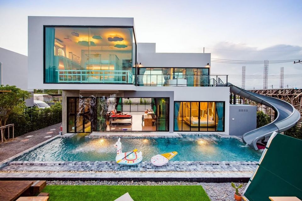 Holiday Sunset Villa IV in Hua Hin. Modern exterior with a large pool, water slide, and illuminated interiors at dusk.
