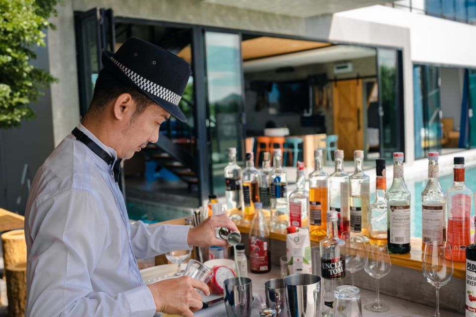 A private bartender in a hat prepares cocktails at a bar with various bottles, overlooking the pool at Holiday Sunset Villa IV, Hua Hin.