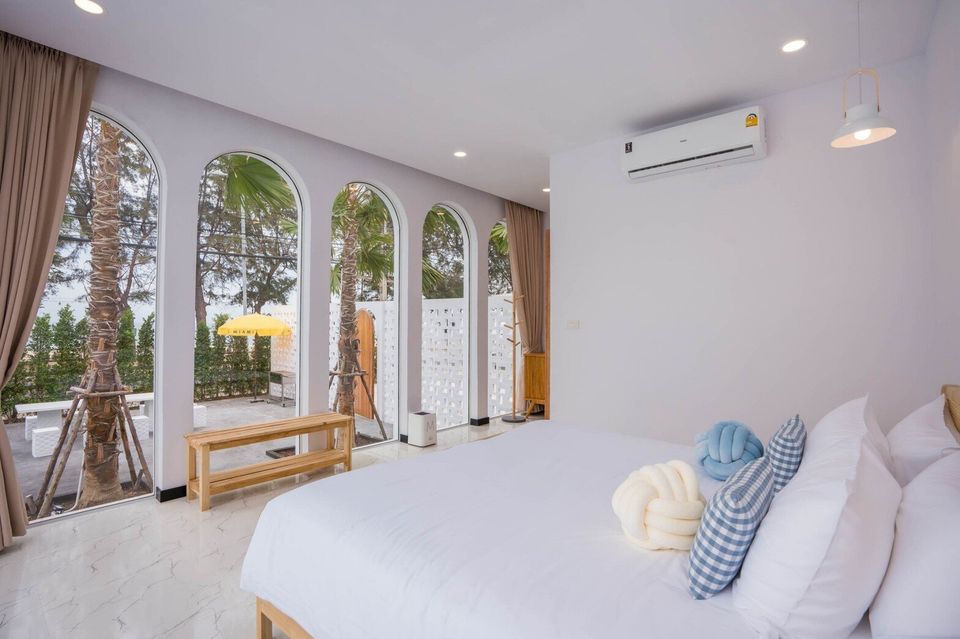 Bright bedroom at Miami Beach VI Pool Villa, Cha-Am, with a comfortable bed and arched windows overlooking palm trees.