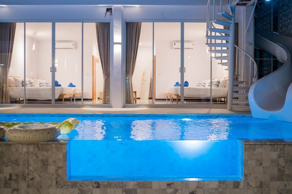 Night view of Miami Beach VI Pool Villa in Cha-Am, featuring a glass-wall pool, water slide, and spiral staircase.
