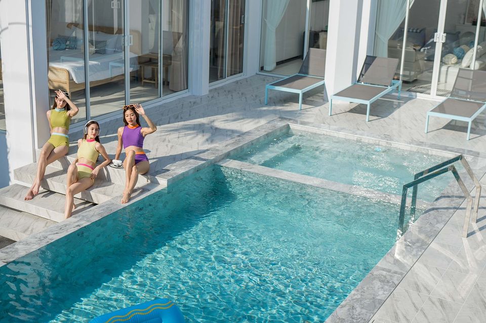Miami Beach VI Pool Villa in Cha Am, Thailand. Three women lounge by the pool with lounge chairs and a view of the bedroom.