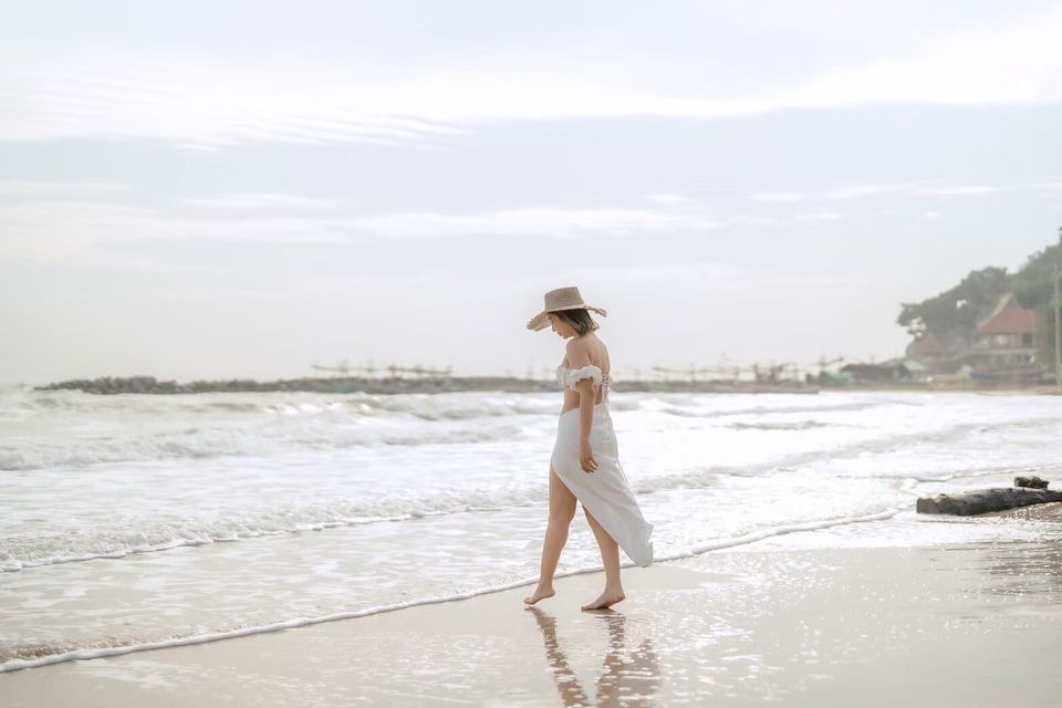 A woman in a straw hat and white dress walks along the sandy beach with waves at Story II Pool Villa Hua-Hin.