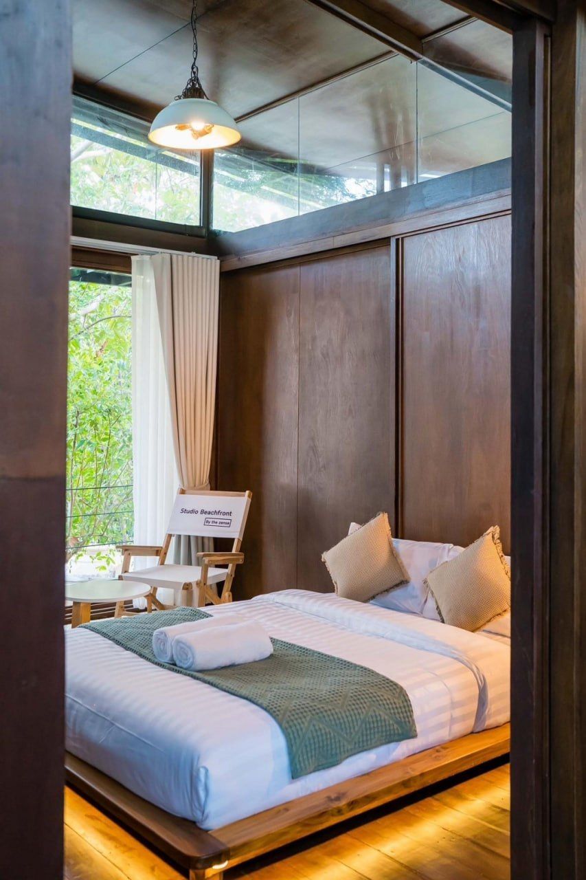 Cozy king-size bed with green throw and two pillows in Studio Beachfront III Pool Villa, Cha-Am. Large window overlooks lush greenery.