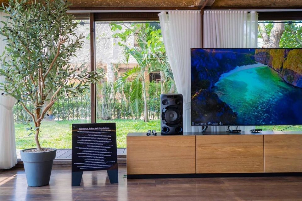 Modern living area with large TV, sound system, and olive tree, offering a lush garden view at Studio Beachfront III Pool Villa, Cha-Am.