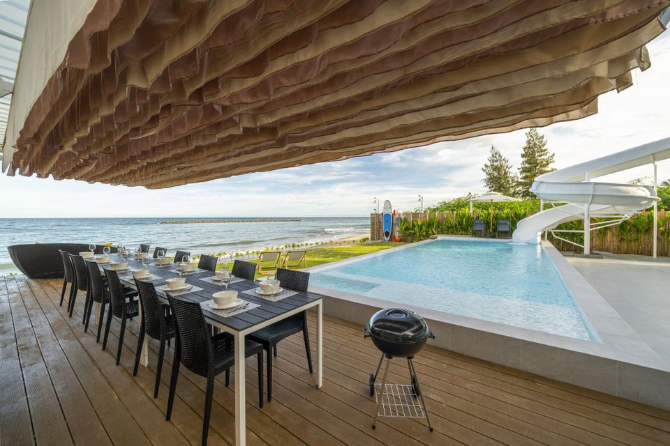 Beachfront outdoor dining table set for guests at Talay Haus X Pool Villa in Cha-Am, with a BBQ grill and pool slide.
