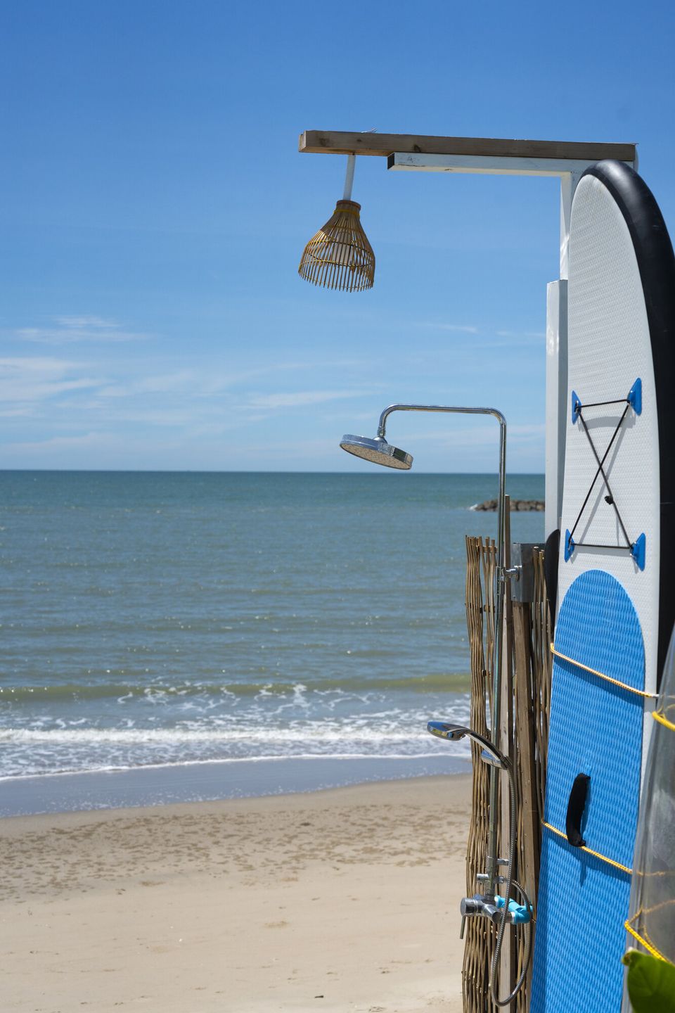 Outdoor beach shower and paddleboard at Talay Haus X Pool Villa in Cha-Am, with the ocean and sandy beach in the background.