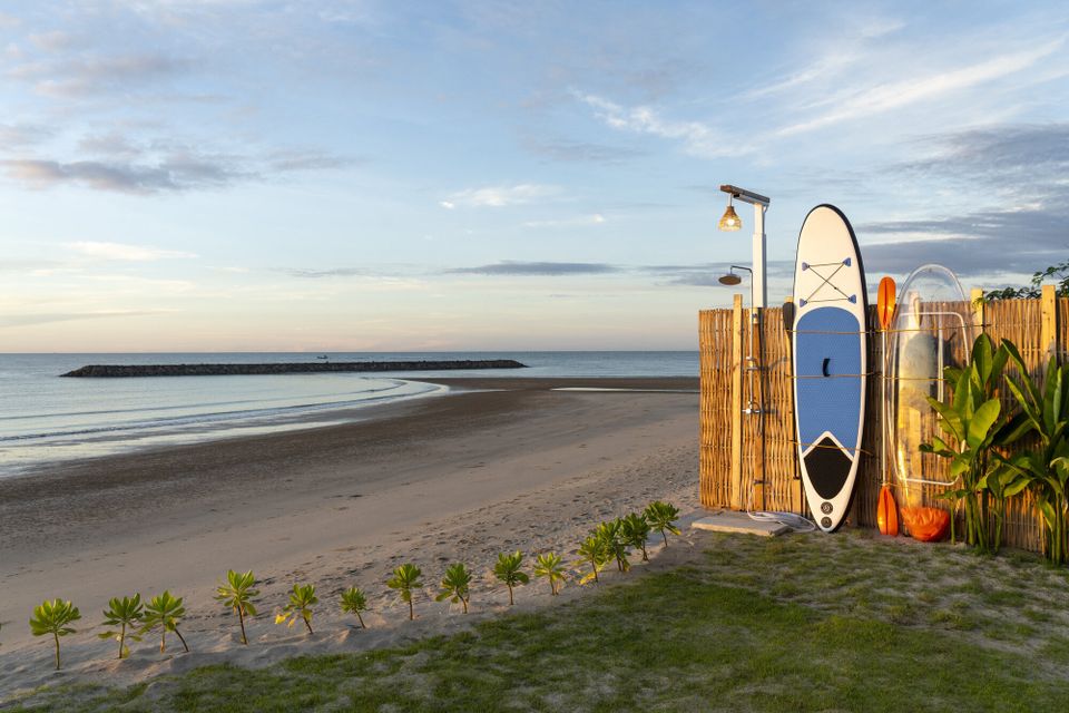 Beachfront outdoor shower and paddleboards at Talay Haus X Pool Villa in Cha-Am, with the ocean and sandy beach at sunset.