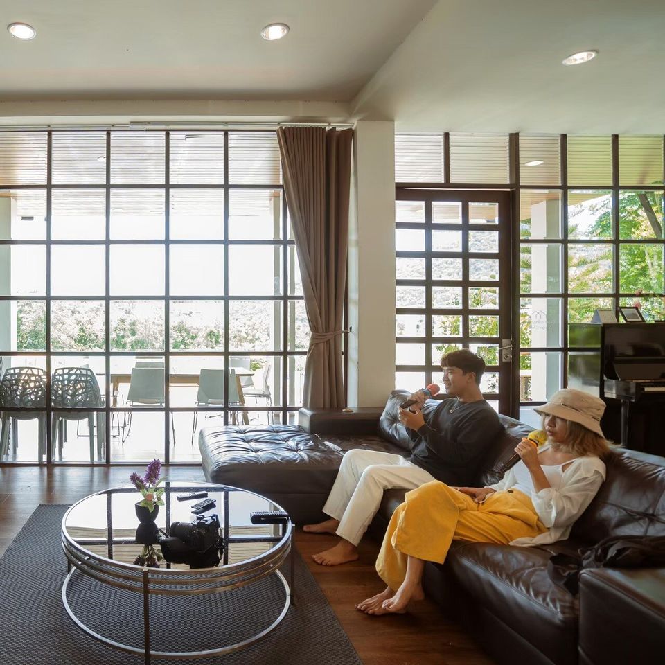 Guests enjoy karaoke in the spacious living room of The Valley Khaoyai V Villa, featuring a comfortable leather sofa and large windows.