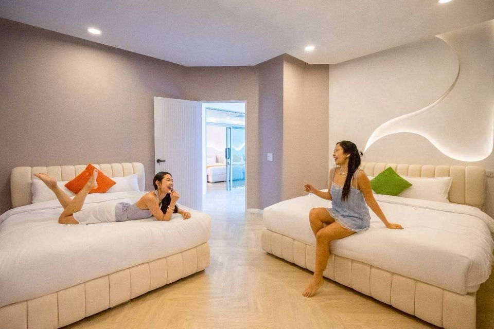 Two women laughing on separate queen beds in a bright bedroom at Tierra V Pool Villa, Pattaya. Modern, comfortable design.